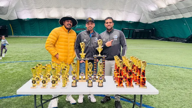 Three adults standing behind a table of trophies at an indoor cricket facility.
